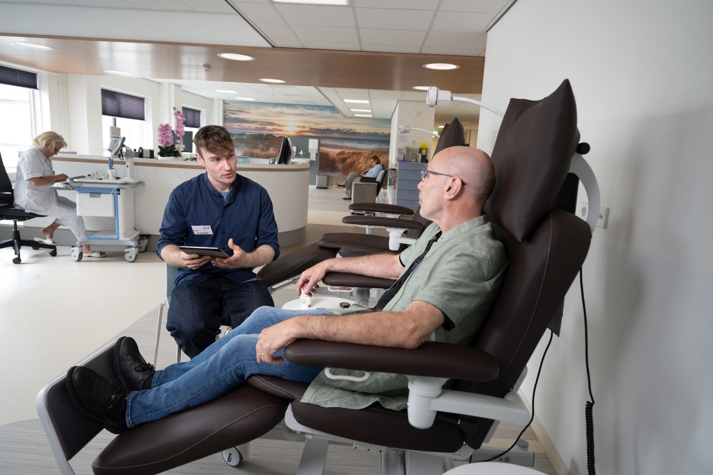 Een vrijwilliger vraagt een patiënt die zit in een behandelstoel op de Dagbehandeling over hoe hij zorg op de afdeling ervaart.